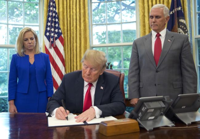 川普总统在白宫椭圆形办公室的坚毅桌上签署行政令，彭斯副总统站在他的左边，国土安全部长尼尔森站在他的右边。(2018年6月20日)