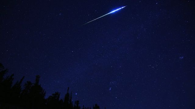 A blue/green glowing meteor crosses the sky. Dark forest visible in the horizon.