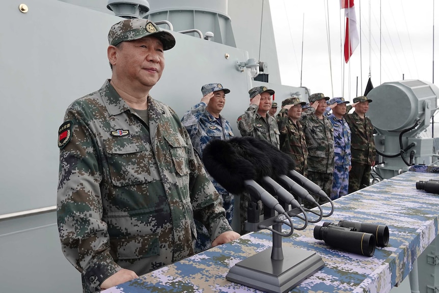 Xi Jinping, in olive, brown and green army fatigues, speaks on a ship after reviewing the naval forces.