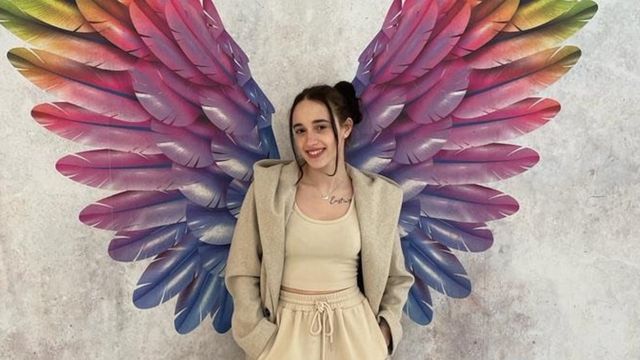 Joana stood against a wall with multi-coloured rainbow wings painted on