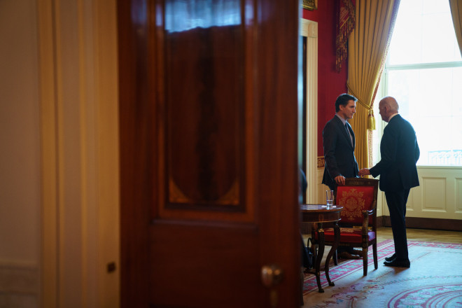 Prime Minister Justin Trudeau and President Joe Biden are standing and speaking. The Prime Minister is leaning against a chair. In the foreground, a door is partially closed.